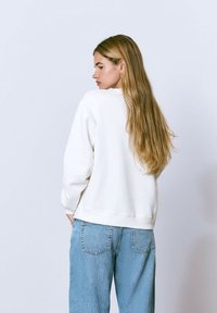 Sudadera blanca con un ajuste casual, mangas largas y puños acanalados, combinada con unos pantalones vaqueros de talle alto de color azul claro. Textura de tela suave.
