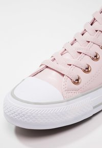 Baskets roses avec embout en caoutchouc blanc, semelle blanche texturée et lacets roses passant par des œillets dorés sur fond blanc.