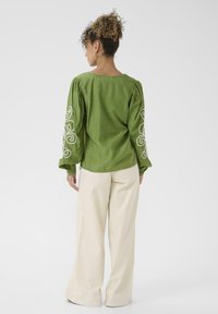 Femme aux cheveux bouclés en chignon portant une blouse à manches longues verte avec de la broderie blanche sur les manches et un pantalon blanc ample, de dos.