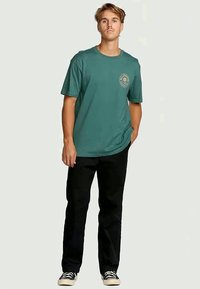 Jonge man die staat in een groen graphic T-shirt, zwarte broek en zwarte sneakers met witte zolen tegen een effen achtergrond.