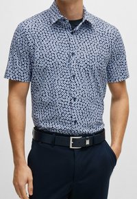 Košile s krátkým rukávem v námořnické modré barvě s bílým květinovým vzorem. Vyrobena z hladkého materiálu, má klasický límec a rovný střih.