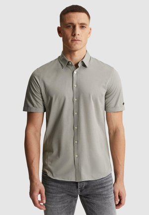 Homme debout, portant une chemise à manches courtes gris clair à boutons et un jean gris délavé sur un fond uni.