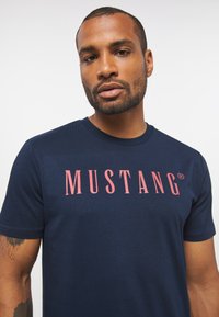 Navy blue cotton t-shirt with short sleeves, featuring a bold red "MUSTANG" logo across the chest. Smooth texture and classic fit.