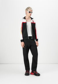 Zwart-witte zip-up jack met rode accenten en "REPLAY" in het geel; gecombineerd met zwarte cargo-broeken en chunky zwart-rode sneakers.