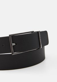Une ceinture en cuir noir avec une boucle rectangulaire à finition métallique, texture lisse et plusieurs trous d'ajustement le long de la sangle.