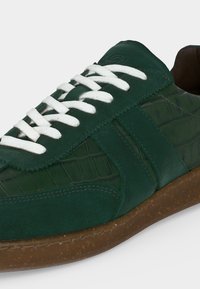 Baskets en cuir vert avec des lacets blancs, des panneaux latéraux texturés et une semelle en caoutchouc marron, vues de l'angle latéral avant.