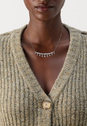 Collier en argent au design courbé ornée de plusieurs petites pierres transparentes, porté par-dessus un cardigan beige texturé avec un détail de bouton.