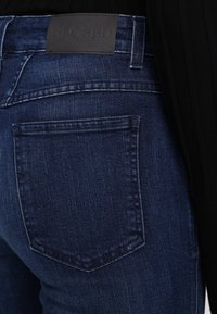 Gros plan sur la poche arrière d'un jean en denim bleu foncé avec des détails cousus et une étiquette de marque en cuir noir sur la ceinture.