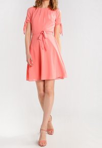 Rosa klänning med kort ärm, cinchad midja och knytdetalj. Utsvängd kjol och mjukt tyg, med knytningar vid de plisserade ärmöppningarna.
