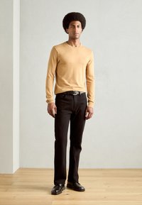 Long-sleeve mustard sweater and black jeans worn by a man, with a black belt and black shoes. Simple design with a relaxed fit.