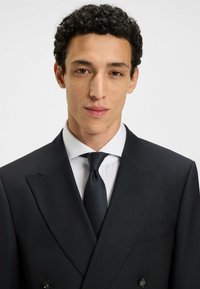 Svart kostym med randigt mönster och dubbelknäppt design, åtföljd av en vit skjorta och en strukturerad mörk slips. Slät tygkvalitet, skräddarsydd passform.