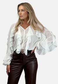 Biała bluzka z falbaniastymi rękawami i przodem, z zapięciem na guziki, zestawiona z brązowymi skórzanymi spodniami i ozdobnym paskiem. Gładki, lekki materiał.