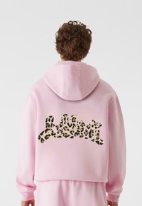 Rožnato kapuco s skrajšanim dizajnom. Zadaj ima črno-bež tekst v vzorcu leoparda. Gladek tekstura tkanine.