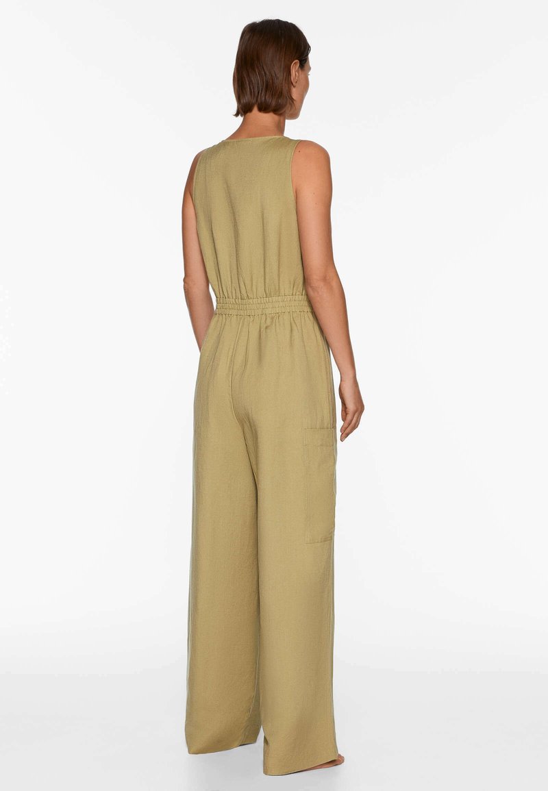 Frau von hinten gesehen, die einen ärmellosen, olivgrünen, weit geschnittenen Jumpsuit mit elastischem Bund und Seitentasche trägt und barfuß vor einem weißen Hintergrund steht.