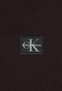Calvin Klein svart merkelapp på dyp burgunder ribbestrikket stoff. Merket har en hvit "CK"-logo og "Calvin Klein"-tekst.