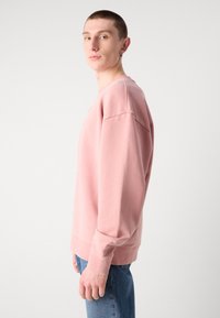 Sudadera rosa de tela suave, con un corte holgado, puños acanalados y escote redondo. Combinada con jeans de mezclilla azules.