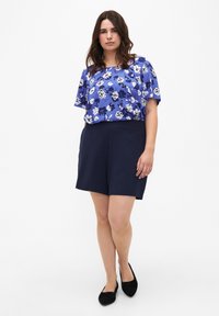 Camiseta corta de flores en azul con flores blancas y negras, combinada con pantalones cortos azul oscuro. Modelo lleva zapatos planos negros con detalle de lazo.