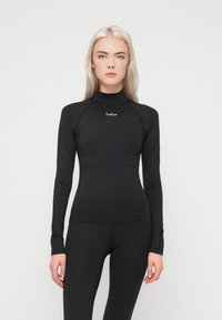 Blond kvinne iført en tettsittende langermet svart treningsgenser og svarte leggings, stående mot en enkel hvit bakgrunn.