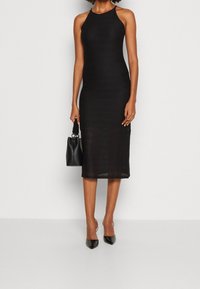 Vestido negro sin mangas con una silueta ajustada y longitud midi. Patrón tejido en todo el vestido; combinado con tacones negros de punta y un bolso pequeño.