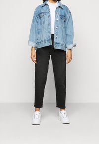 Person klädd i en ljusblå denimjacka, vit t-shirt, svarta midjehöga byxor och vita sneakers som står framför en enfärgad bakgrund.