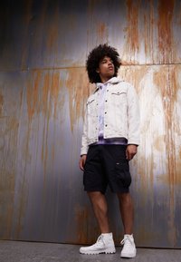 Giubbotto di denim bianco sopra un hoodie tie-dye, pantaloni cargo neri e stivali bianchi. Lo sfondo presenta una superficie metallica arrugginita.