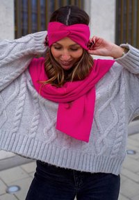 Maglione grigio a maglia grosso con una vestibilità ampia, abbinato a una fascia per capelli annodata di un brillante colore rosa e a una sciarpa coordinata, in un contesto informale all'aperto.