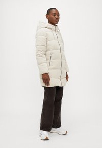 Cremefarvet pufferjakke med quiltet design, frontlynlås og hætte. Parret med brune cordbukser og hvide sneakers.