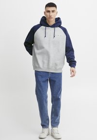 Grijze hoodie met marineblauwe mouwen en capuchon. Bevat een frontzak, trekkoord en een klein logo op de borst. Gecombineerd met blauwe jeans en witte sneakers.