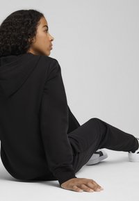 Sweatshirt noir avec capuche et pantalon assorti. Tissu doux, coupe décontractée, poignets côtelés, et des baskets blanches qui contrastent avec la tenue.