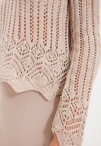 Beżowy sweter z dzianiny o ażurowym wzorze, z pionowymi paskami i skomplikowanymi geometrycznymi wzorami wzdłuż dolnego brzegu. Teksturowany, lekki materiał.