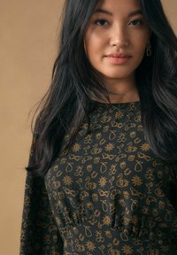 Sort stoftop med guldmønstre af forskellige objekter som frugter, dyr og solmotiver; indsamlet design ved taljen, lange ærmer.