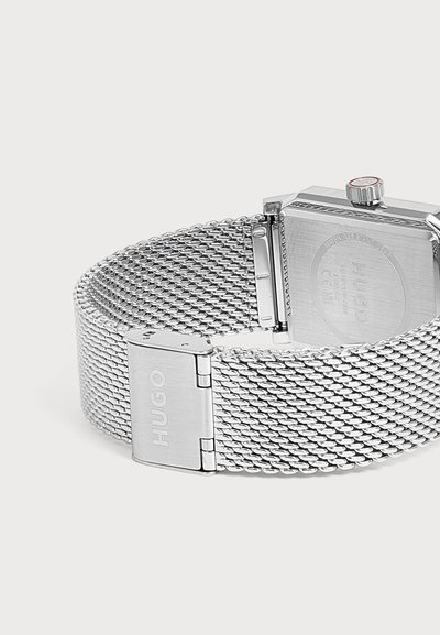 Bracelet de montre en métal maille argenté avec une boucle gravée "HUGO" et le dos d'un boîtier de montre carré avec une couronne à pointe rouge.