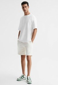 T-shirt bianca oversize con maniche corte, abbinata a pantaloni corti color crema e sneaker bianche con accenti verdi. Tessuto morbido e vestibilità rilassata.