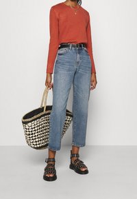 Maglione a maniche lunghe color ruggine, jeans dritti a vita alta blu, sandali neri con borchie e una borsa di paglia intrecciata con accenti neri.