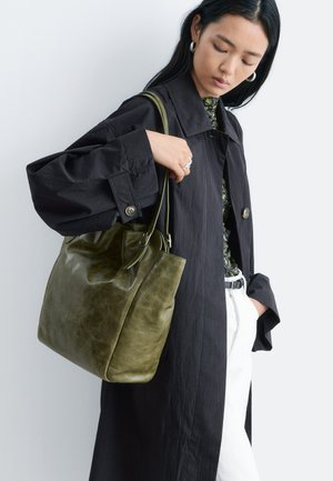 Mujer con abrigo negro y pantalones blancos sostiene un gran bolso tote de cuero verde oliva al hombro contra un fondo liso.