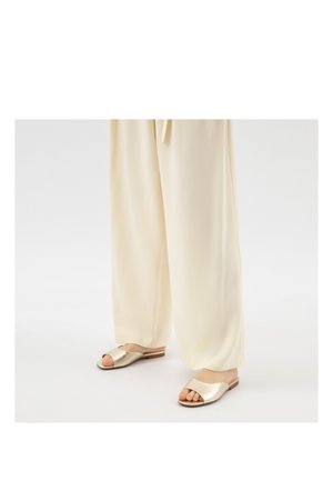 Nohy v volných krémových kalhotách a kovově zlatých sandálech s otevřenou špičkou a plochou podrážkou stojí na bílém podlaze.