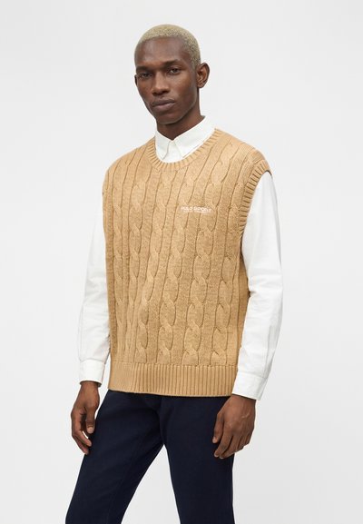 Άνδρας με κοντά ξανθά μαλλιά φοράει μπεζ αμάνικο πουλόβερ με πλεκτό σχέδιο πάνω από λευκό πουκάμισο με γιακά και σκούρο παντελόνι, στέκεται μπροστά σε λευκό φόντο.