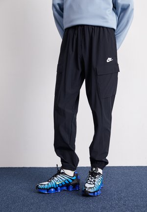 Pantalones cargo negros con cinturilla elástica y bolsillos, combinados con zapatillas Nike multicolores que tienen una suela azul y un diseño detallado en la parte superior.