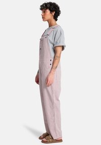 Ριγέ ολόσωμες φόρμες από βαμβάκι κόκκινες και λευκές, με ρυθμιζόμενους ιμάντες, πλαϊνές τσέπες και λεπτομέρειες με κουμπιά, συνδυασμένες με γκρι κοντομάνικο πουκάμισο.