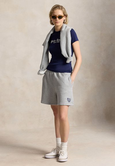 Femme portant un t-shirt bleu marine, un short gris avec logo, un pull gris posé sur les épaules, des chaussettes blanches, des baskets blanches et des lunettes de soleil, debout devant un fond uni.