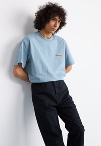Lys blå bomulds T-shirt med korte ærmer og rund halsudskæring, med et lille flerfarvet logo på brystet, parret med mørke bukser.