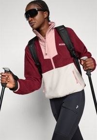 Burgunder- og rosa fleecepullover med lynlås. Hvit nedre del, to sidesyninger. Modell som bærer en svart ryggsekk og holder på med stavene.