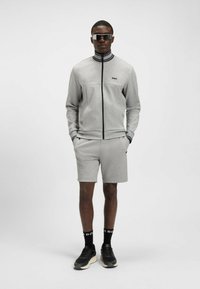 Giacca grigia con zip e colletto arrotondato, abbinata a pantaloni corti coordinati. Gli accessori includono occhiali da sole neri e scarpe sportive.