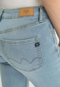 Jean en denim bleu clair avec une poche arrière ornée d'un design en forme de cœur brodé et une étiquette de marque en cuir beige près de la taille.
