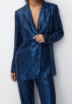 Femme portant un blazer bleu profond à sequins et un pantalon assorti, tenant d'une main le revers du blazer sur un fond uni.
