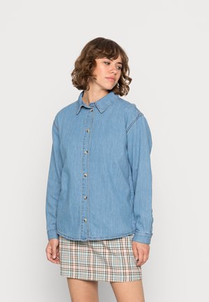 Camisa de jeans azul-claro com mangas longas, colarinho e botões frontais. Detalhe de costura notável ao longo dos ombros.