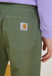 Zbliżenie na tylną kieszeń oliwkowych spodni z naszywką logo Carhartt, obok dłoni i części lawendowego rękawa.