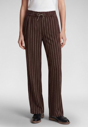 Woman wearing brown drawstring pants with white vertical stripes and black loafers, standing against a plain gray background.