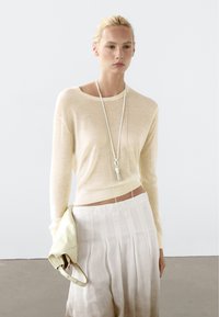 Beige lichtgewicht trui met een ronde hals, gecombineerd met een geplooide crèmekleurige rok. Accessoires omvatten een lange hangerketting en een lichte clutch.