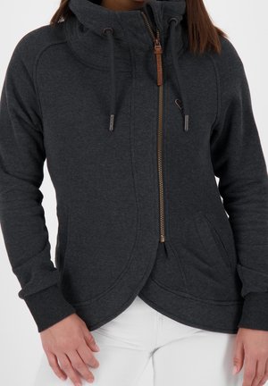 Femme portant un sweat à capuche zippé gris foncé avec col haut et cordons, associée à un pantalon blanc, debout les mains détendues.